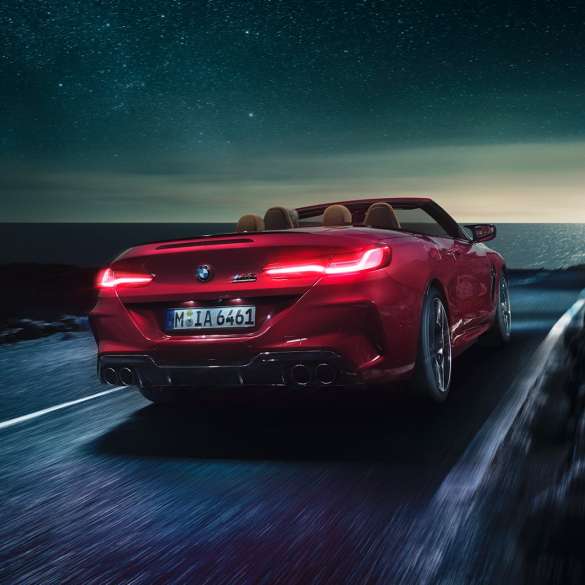 BMW M8 Competition Cabrio, arkadan görünüm. Metalik Kırmızı/Motegi BMW M8 Competition Cabrio sahil yolunda açık softtop tavanla gece sürüşü sırasında, arkadan görünüm.