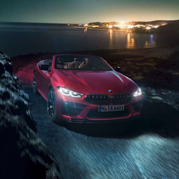 BMW M8 Competition Cabrio, önden görünüm. Metalik Kırmızı/Motegi BMW M8 Competition Cabrio sahil yolunda açık softtop tavanla gece sürüşü sırasında, önden görünüm.