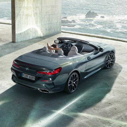 BMW M850i xDrive Cabrio, kuş bakışı görünüm. Açık softtop ile Metalik Gri/Dravit BMW M850i xDrive Cabrio, okyanus kenarında beton bir bina içinde park halinde, kuş bakışı görünüm.