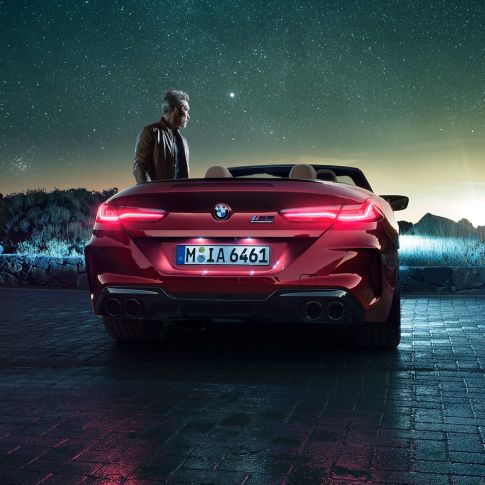 BMW M8 Competition Cabrio, arkadan görünüm. Açık softtop ile Metalik Kırmızı/Motegi BMW M8 Competition Cabrio, yanında duran bir kişiyle yıldızlı gökyüzü altında arkadan görünüm.