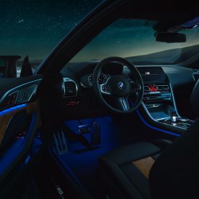 BMW M8 Competition Coupé’nin iç tasarımı. BMW M8 Competition Coupé, M deri direksiyon ve mavi ortam aydınlatmasıyla sürücü kapısı açık iç tasarımın gece görünümü.