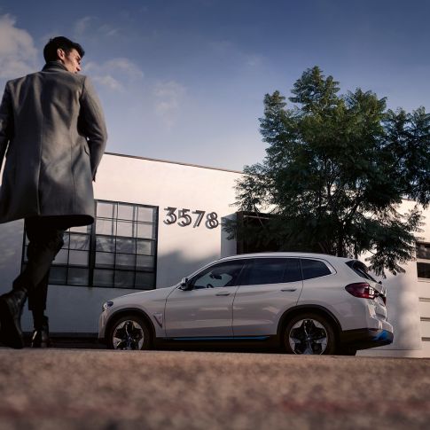 BMW iX3 G08 bina önünde adam ve yandan görünüm BMW iX3 G08 Metalik Beyaz/Mineral bina önünde adam ve yandan görünüm