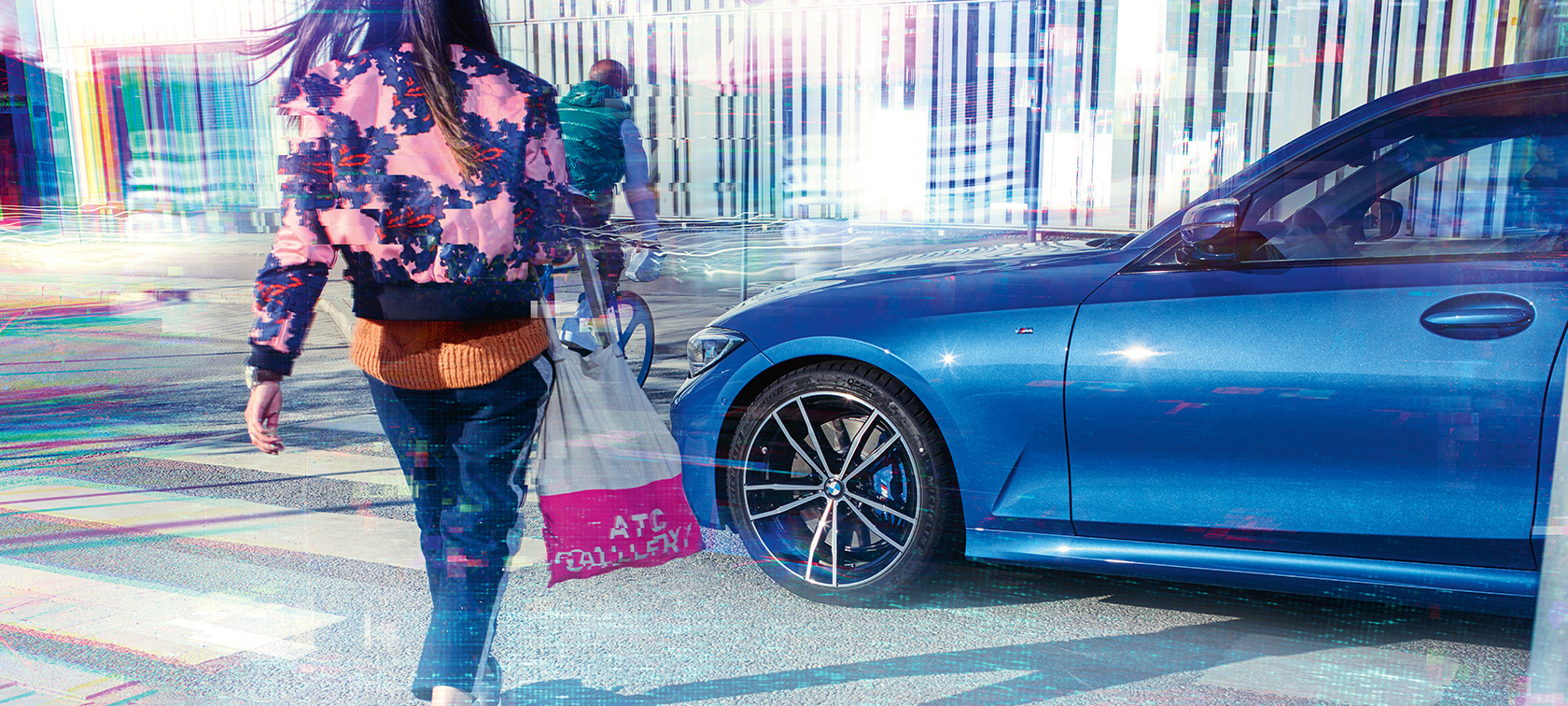 Yeni BMW 3 Serisi Sedan, yandan çekim. Yeni BMW 3 Serisi Sedan’ın yolcularla birlikte şehir manzarası önünde asfalt üzerinde yandan görünümü.