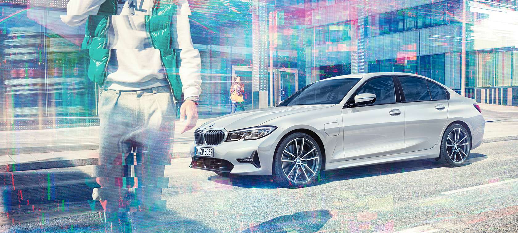 Yeni BMW 3 Serisi Sedan, BMW 330e yandan görünümü. Yürüyen yolcularla birlikte şehir içinde camlı bir binanın önünde hareketsiz halde BMW 330e iPerformance Sedan’ın yandan görünümü.