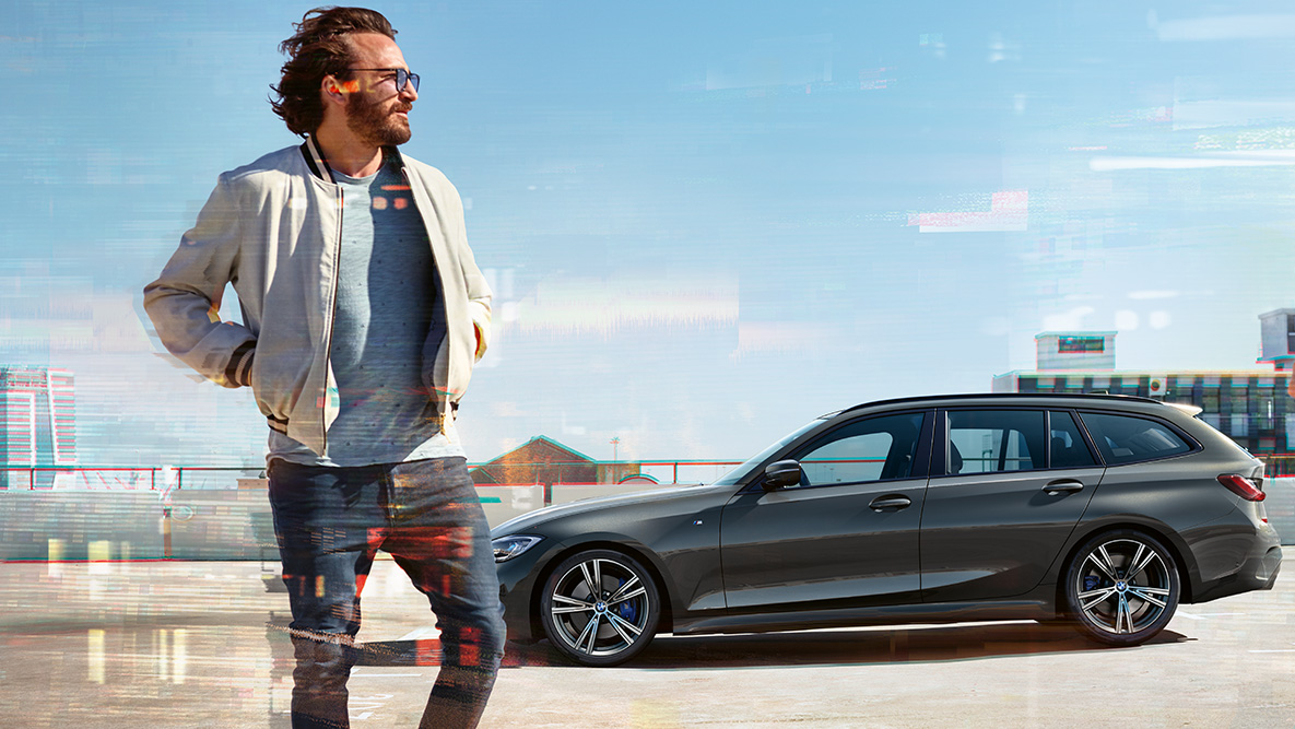BMW 3 Serisi Touring (G21), tasarım, ön çaprazdan görünüm BMW 3 Serisi Touring (G21), Metalik Gri/Dravite, kır manzarası önünde köprüden geçerken çekilmiş ön çaprazdan görünüm