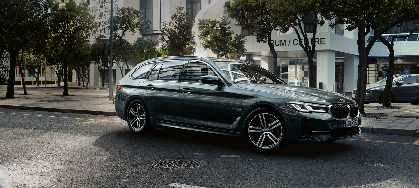 BMW 5 Serisi Touring G31, Metalik Gri/Sophisto, şehrin eteklerinde sürüş sırasında ön çaprazdan görünüm