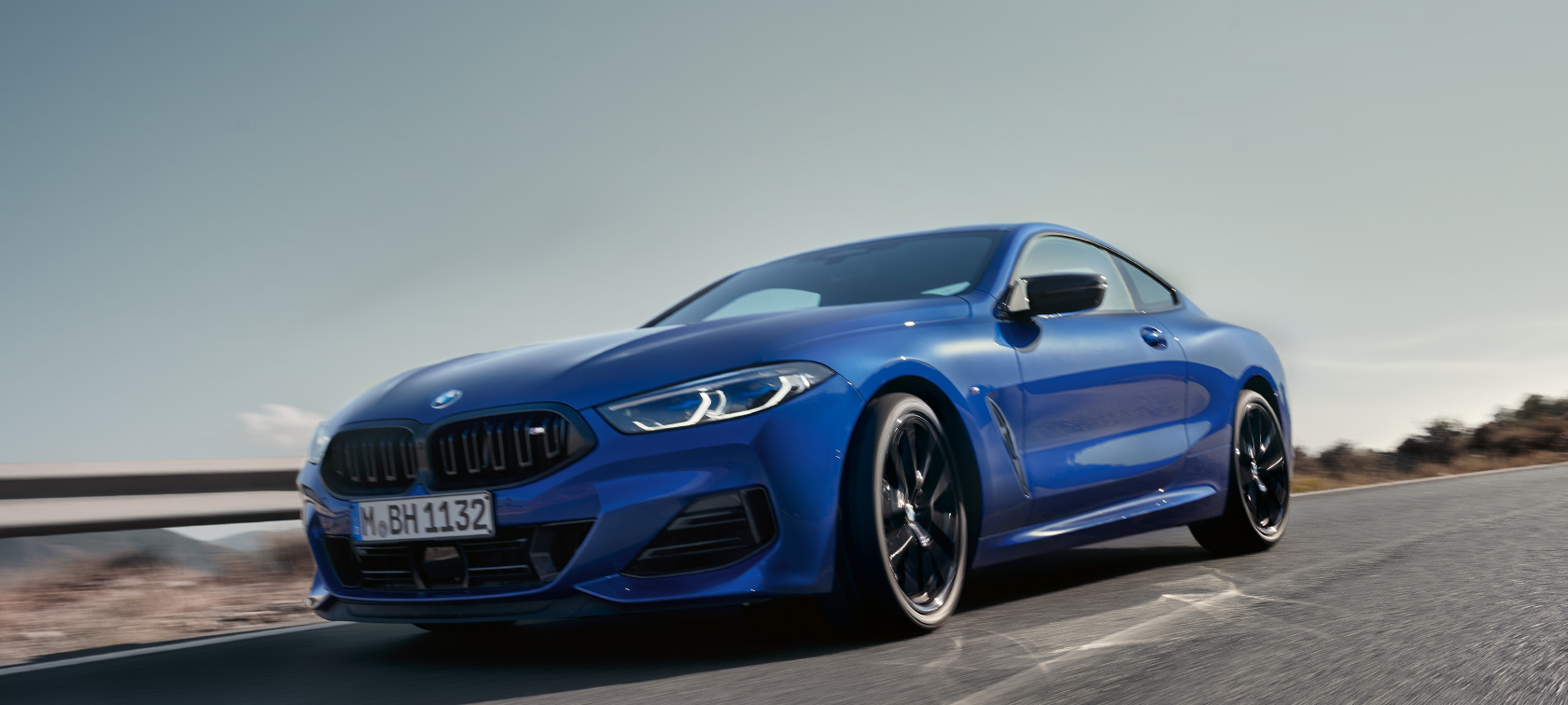 BMW 8 Serisi Coupé G15 Metalik Mavi/M Portimao renginde, alçak çekim önden görünüm BMW 8 Serisi Coupé G15 Metalik Mavi/M Portimao renginde, arka planda tepelerin bulunduğu manzara önünde kırsal yolda sürüş sırasında alçak çekim önden görünüm