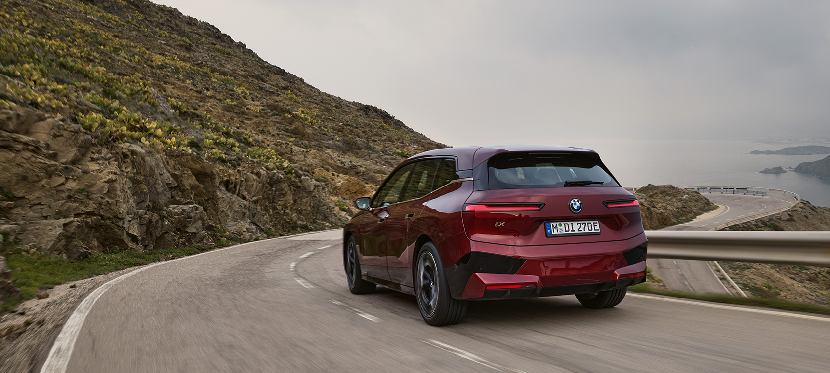 BMW iX i20 elektrikli SUV, BMW iX xDrive50, sahil yolunda sürüş BMW iX i20 elektrikli SUV, BMW iX xDrive50, Metalik Kırmızı/Aventurin, sahil yolunda sürüş sırasında arkadan görünüm