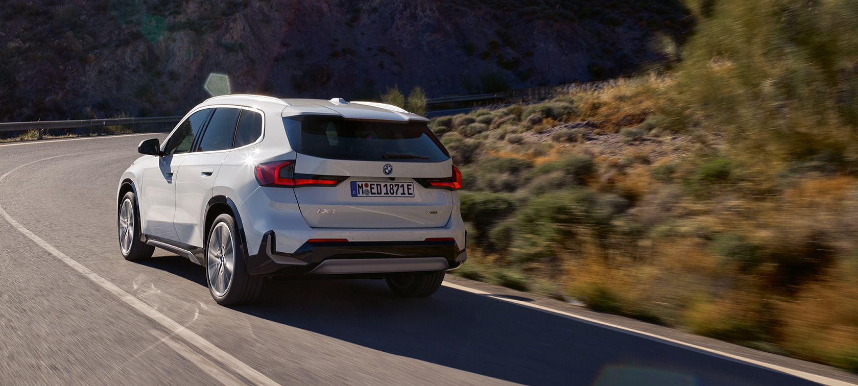 BMW iX1 U11 BEV, Öne çıkan sürüş dinamikleri videosu BMW iX1 U11 BEV, Metalik Beyaz/Mineral, Dağ yolunda sürüş sırasında arka çaprazdan görünüm, Öne çıkan sürüş dinamikleri videosu
