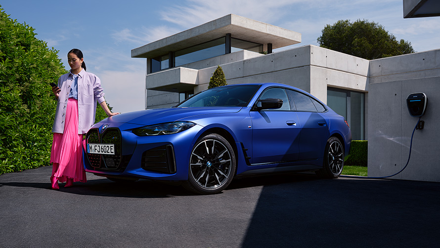 BMW i4 M50 G26, BMW Individual Metalik Mat Mavi/Portimao, evin önünde bir kadın ile ön çaprazdan görünüm