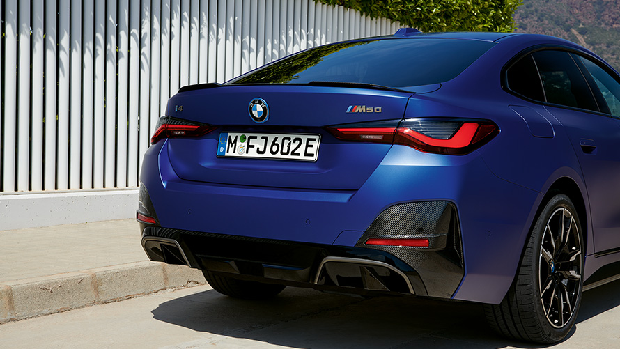 BMW i4 M50 G26, BMW Individual Metalik Mat Mavi/Portimao, beyaz bir çitin önünde arka çaprazdan görünüm