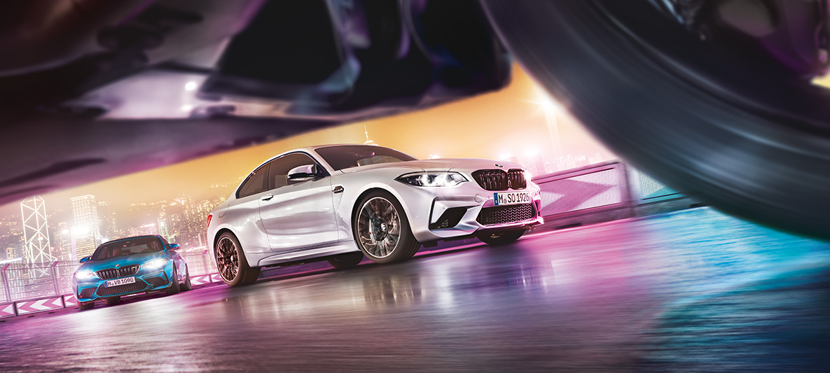 Yeni BMW M2 Competition otomobili asfalt yol üzerinde. İki Yeni BMW M2 Competition otomobil gece şehir manzarası önünde yarış pistinde drift yapıyor.