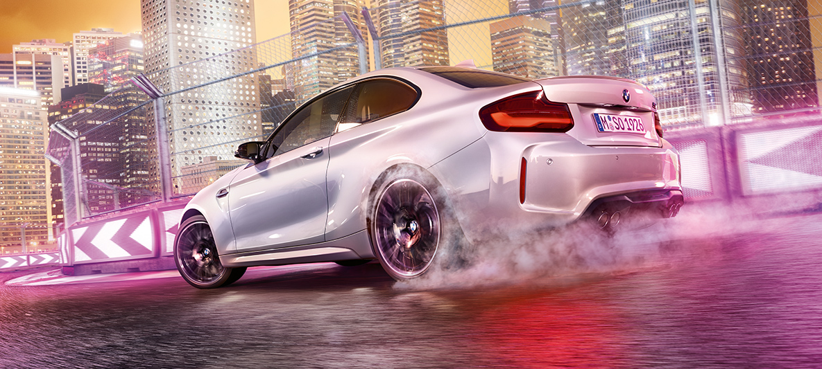 Yeni BMW M2 Competition hızlanıyor Beyaz renkte Yeni BMW M2 Competition gece şehir manzarası önünde yarış pistinde drift yapıyor.