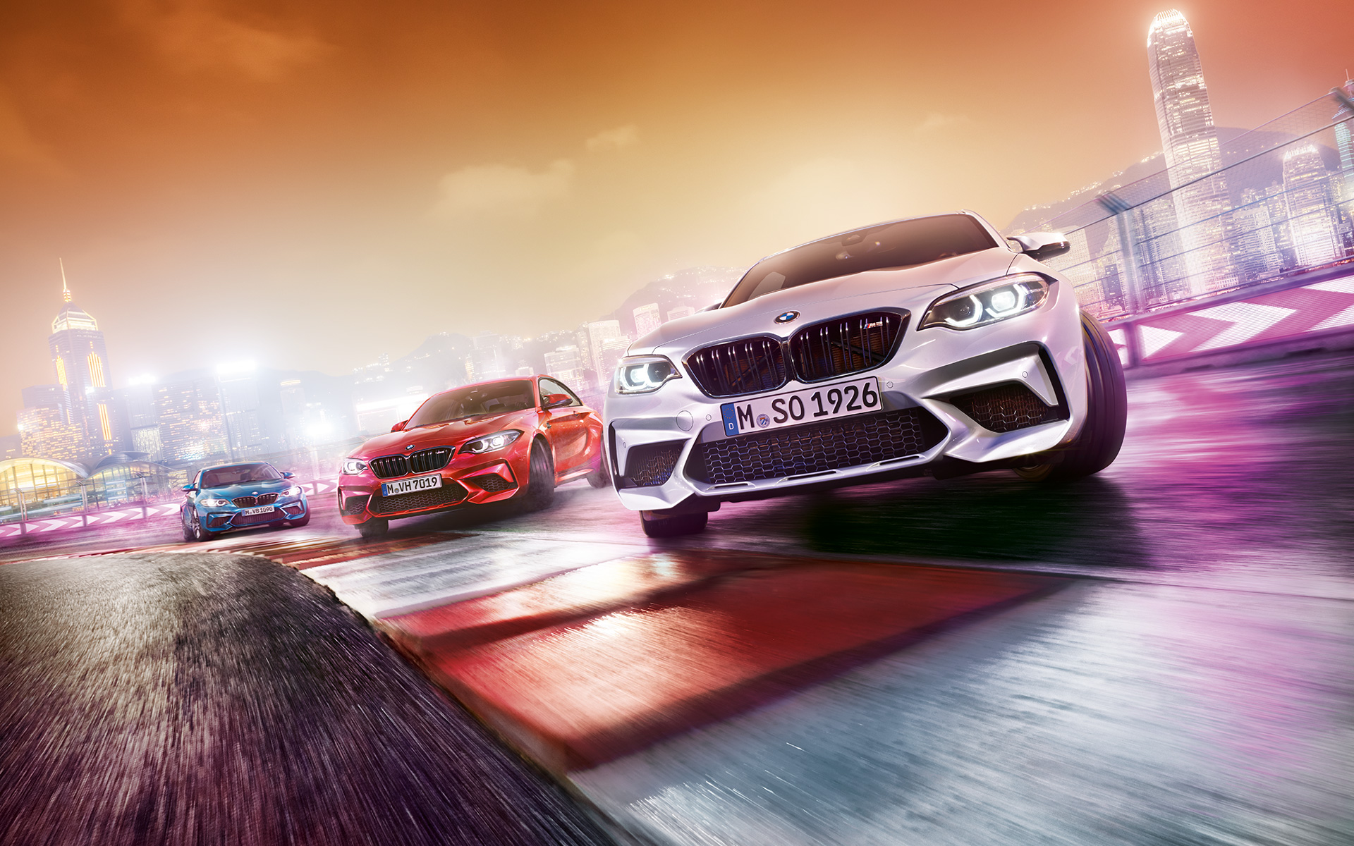 Yeni BMW M2 Competition hareket halinde Üç adet Yeni BMW M2 Competition otomobili gece şehir manzarası önünde yarış pistinde drift yapıyor.