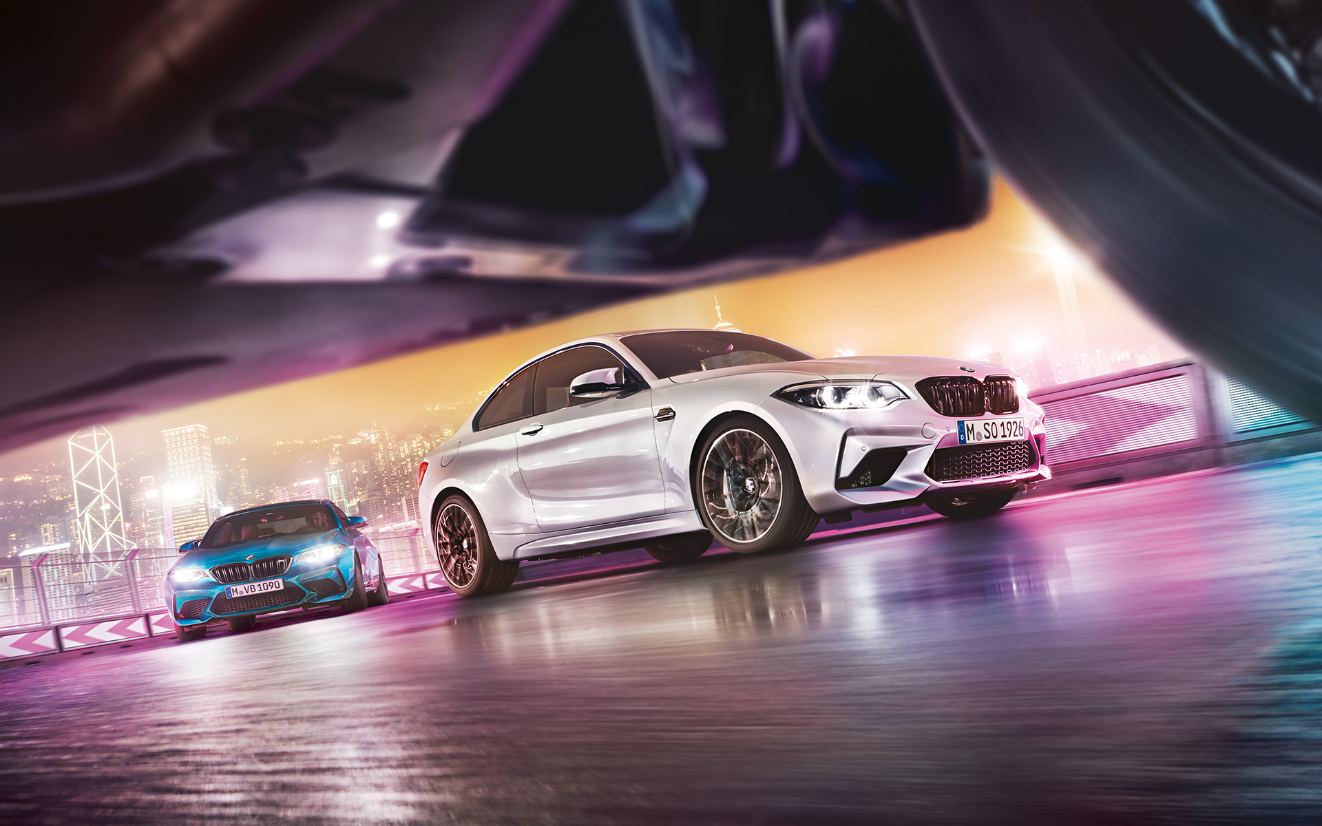 Yeni BMW M2 Competition yansıtıcı asfalt üzerinde İki adet Yeni BMW M2 Competition otomobil sokak seviyesinde gece şehir manzarası önünde yarış pistinde drift yapıyor.