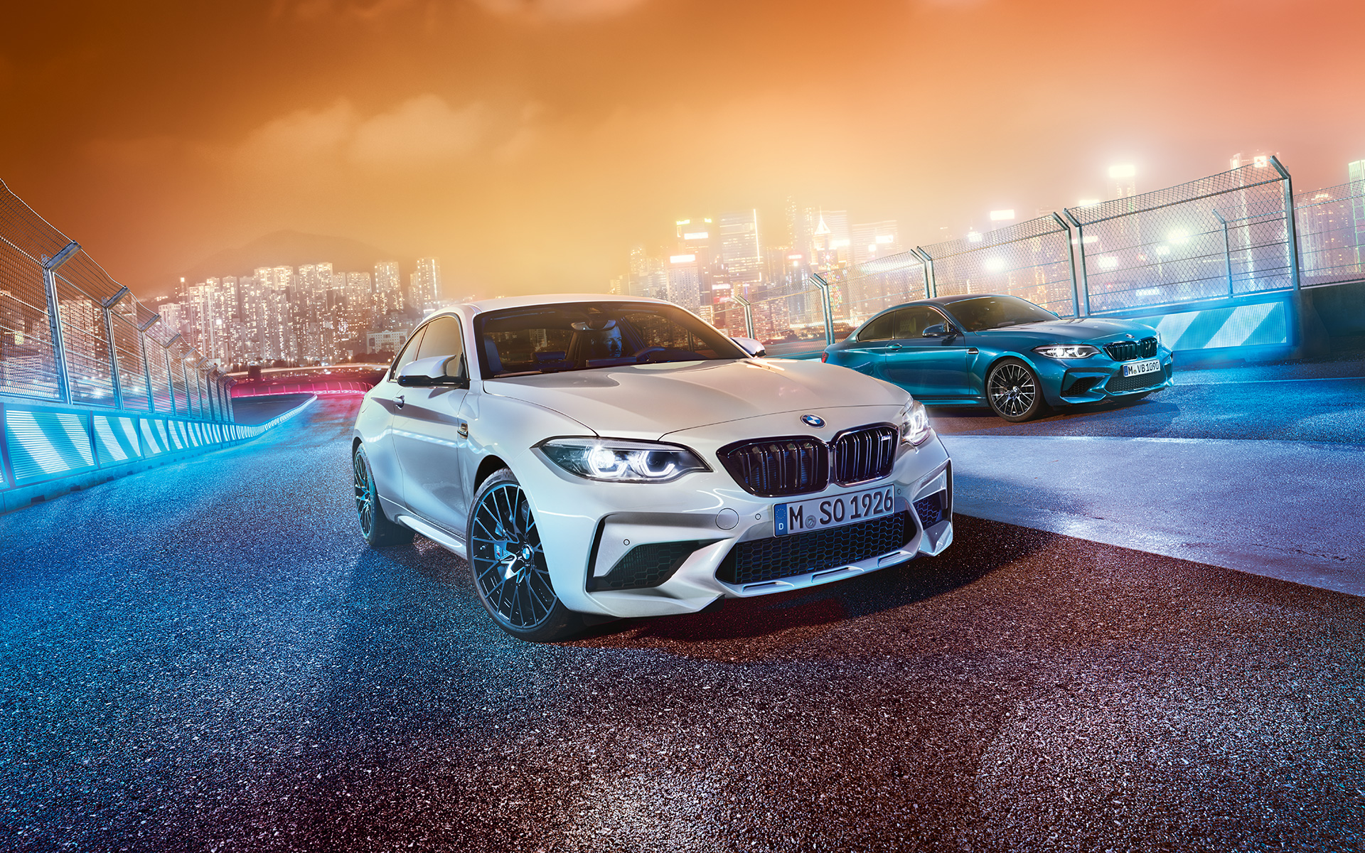 Yarış pistinde iki Yeni BMW M2 Competition Beyaz ve mavi renkte Yeni BMW M2 Competition ile gece şehir manzarası - çapraz ön görünüm.