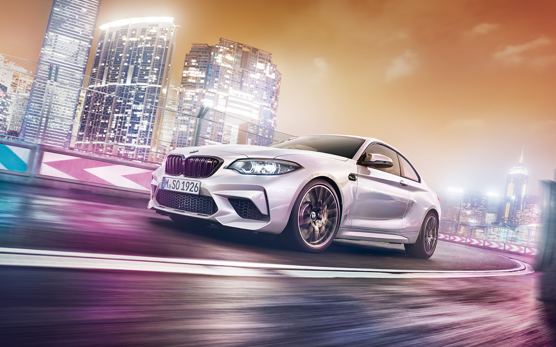 Ufuk çizgisinde beyaz renkte Yeni BMW M2 Competition Beyaz renkte Yeni BMW M2 Competition gece şehir manzarası önünde yarış pistinde ilerliyor.