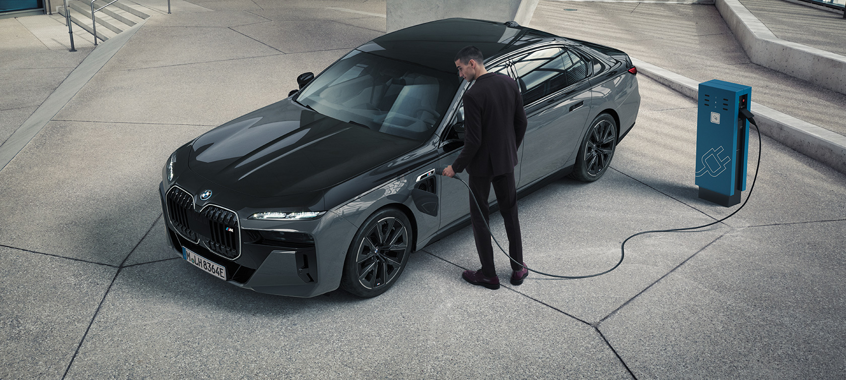 BMW M760e xDrive Sedan G70 M Performance, BMW Individual Metalik Siyah/Sapphire BMW Individual Metalik Gri/Dravite iki tonlu gövde rengi, şarj istasyonunda şarj kablosu tutan bir adam ile ön çaprazdan görünüm