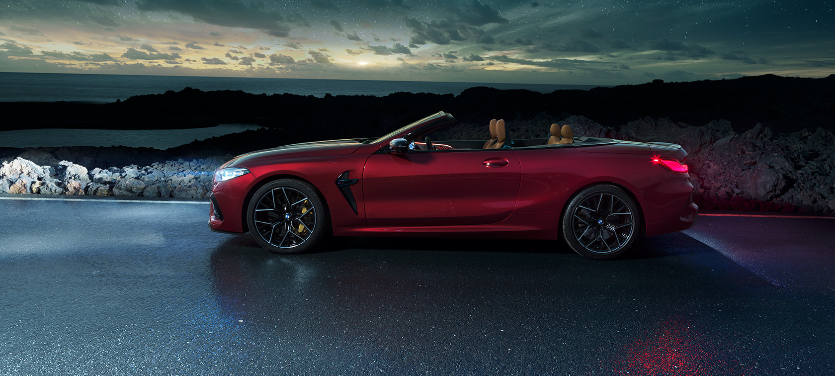 BMW M8 Competition Cabrio, yandan görünüm. Açık softtop ile metalik Kırmızı/Motegi BMW M8 Competition Cabrio, gece yıldızlı gökyüzü altında sahil yolunda, yandan görünüm.