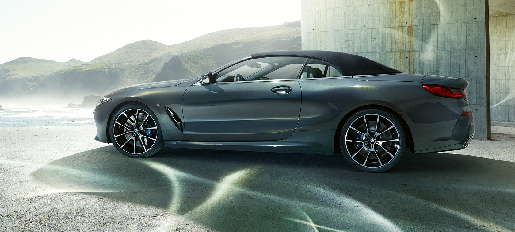 BMW M850i xDrive Cabrio, yandan görünüm. Kapalı softtop ile Metalik Gri/Dravit BMW M850i xDrive Cabrio, arka planda tepe manzarasıyla yandan görünüm.