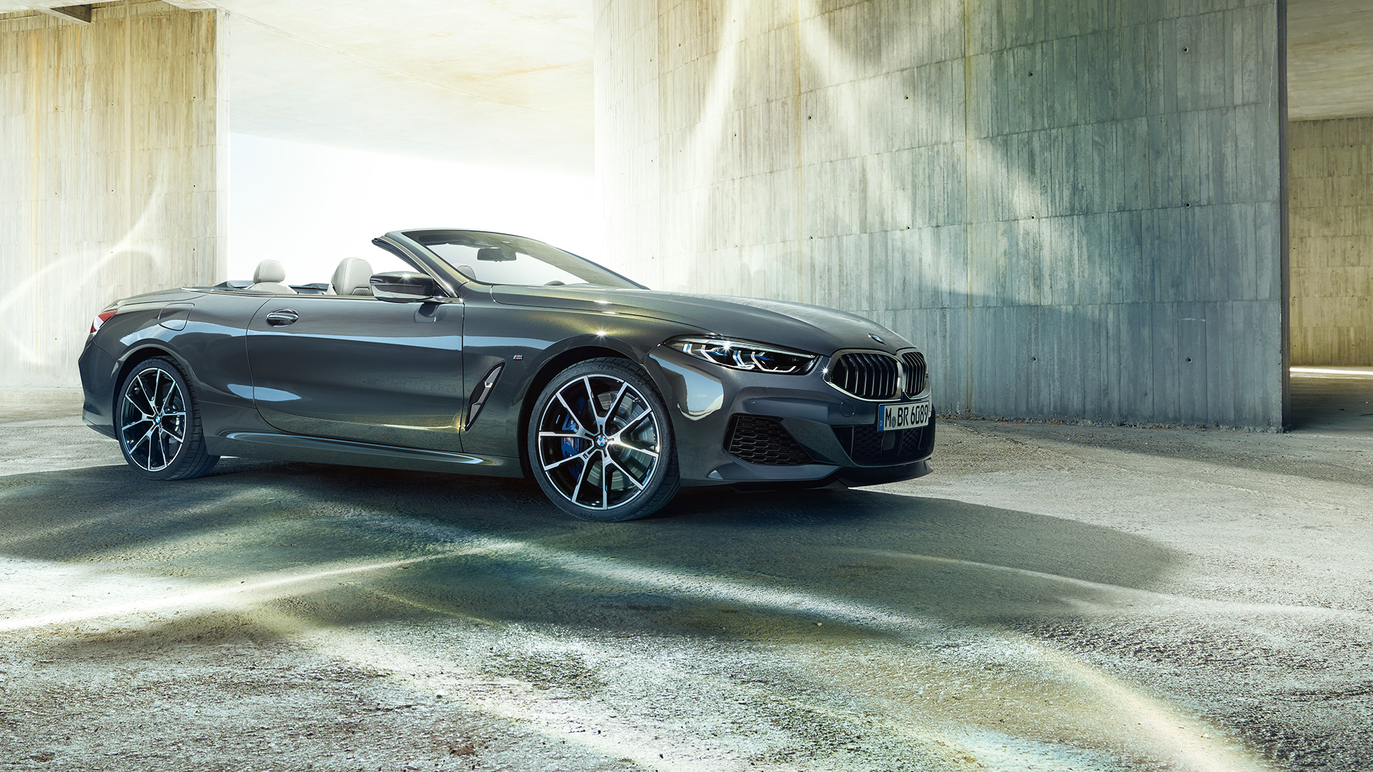 BMW M850i xDrive Cabrio, yandan görünüm. Açık softtop ile Metalik Gri/Dravit BMW M850i xDrive Cabrio, beton bina önünde, yandan görünüm.