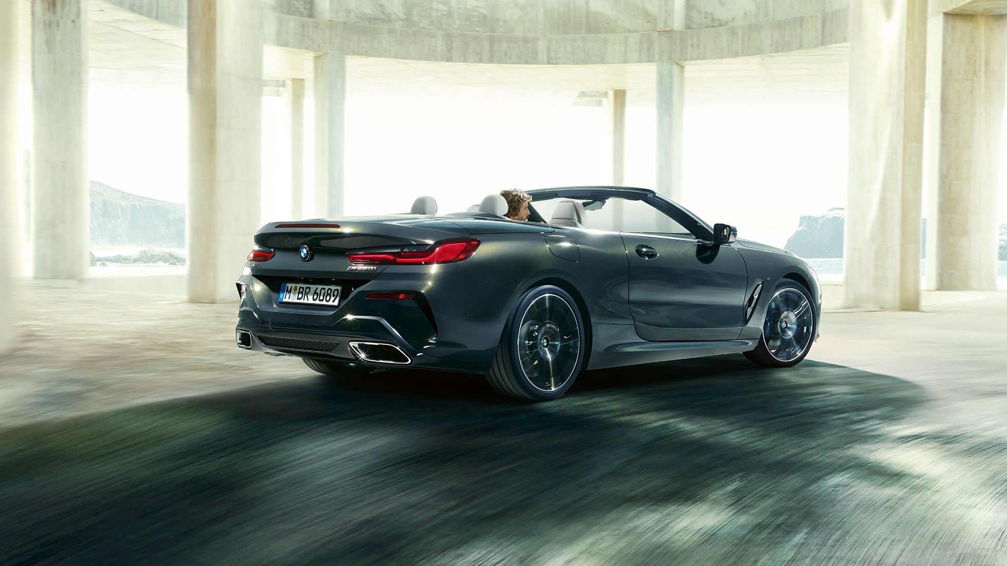 BMW M850i xDrive Cabrio, arkadan görünüm. Açık softtop ile metalik Gri/Dravit BMW M850i xDrive, beton bina yanından geçerken, arkadan görünüm.