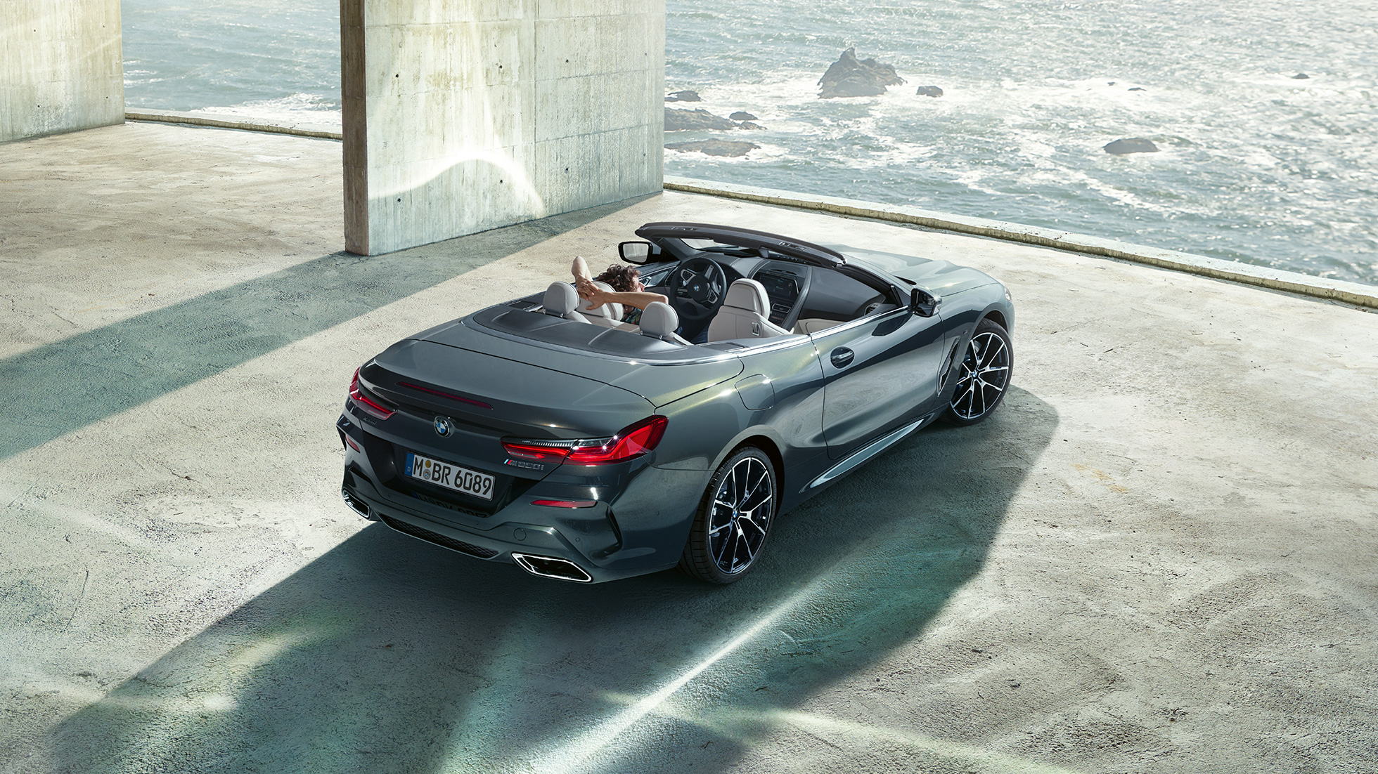 BMW M850i xDrive Cabrio, kuş bakışı görünüm. Açık softtop ile Metalik Gri/Dravit BMW M850i xDrive Cabrio, okyanus kenarında beton bir bina içinde park halinde, kuş bakışı görünüm.