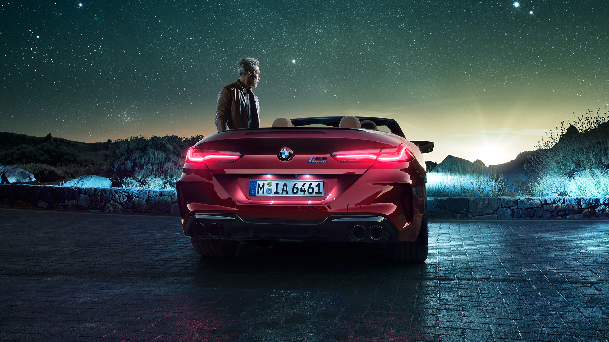BMW M8 Competition Cabrio, arkadan görünüm. Açık softtop ile Metalik Kırmızı/Motegi BMW M8 Competition Cabrio, yanında duran bir kişiyle yıldızlı gökyüzü altında arkadan görünüm.