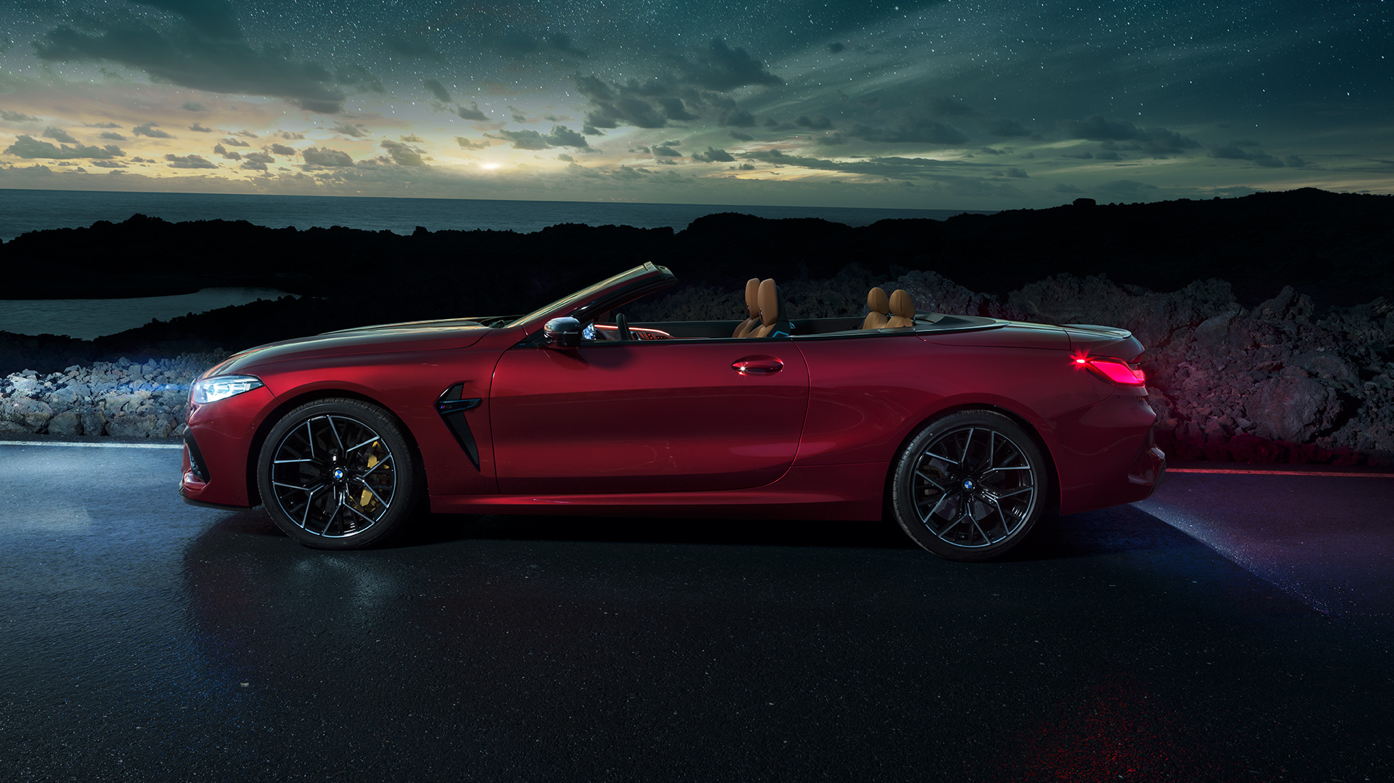 BMW M8 Competition Cabrio, yandan görünüm. Açık softtop ile Metalik Kırmızı/Motegi BMW M8 Competition Cabrio, yıldızlı gökyüzü altında yandan görünüm.