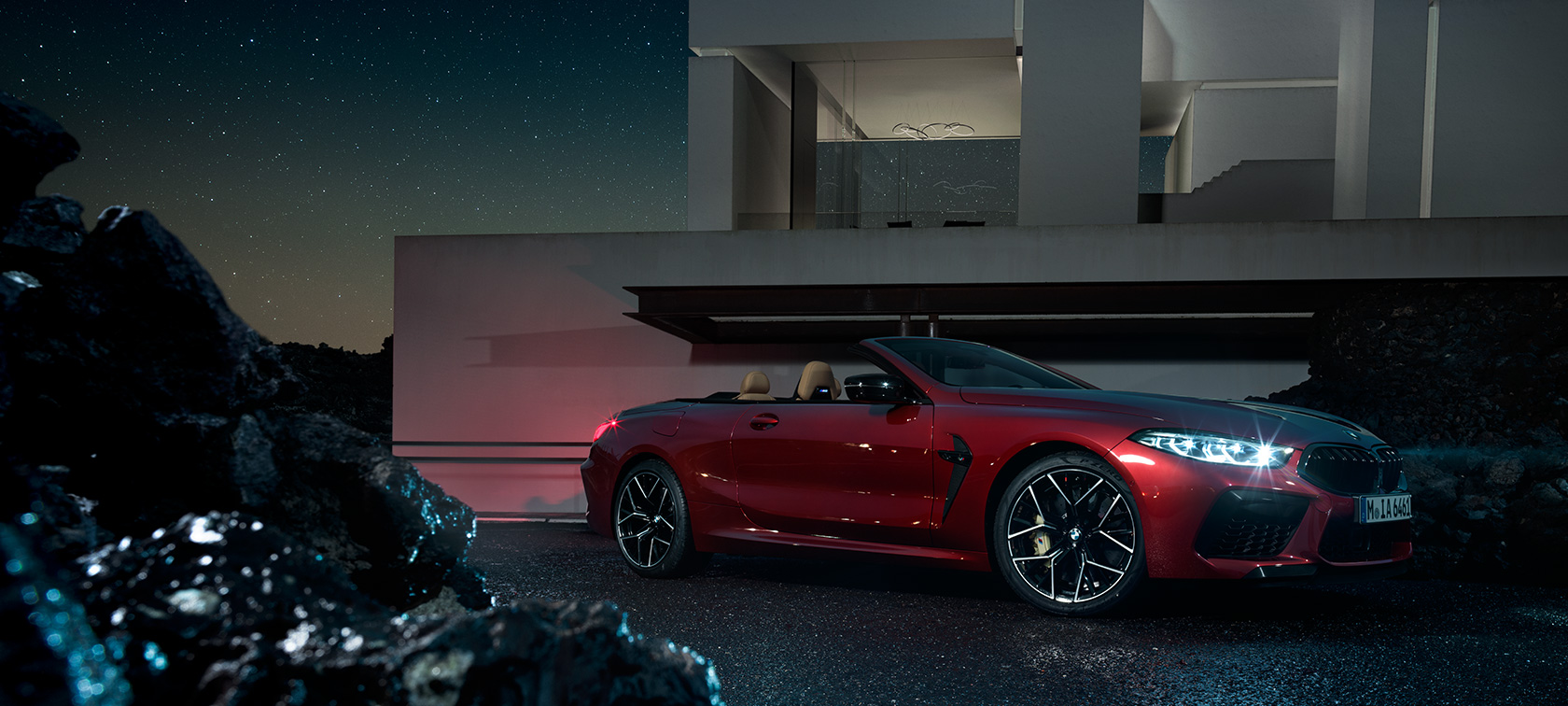 BMW M8 Competition Cabrio, yandan görünüm. Metalik Kırmızı/Motegi renginde BMW M8 Competition Cabrio gece bir villanın önünde, yandan görünüm.