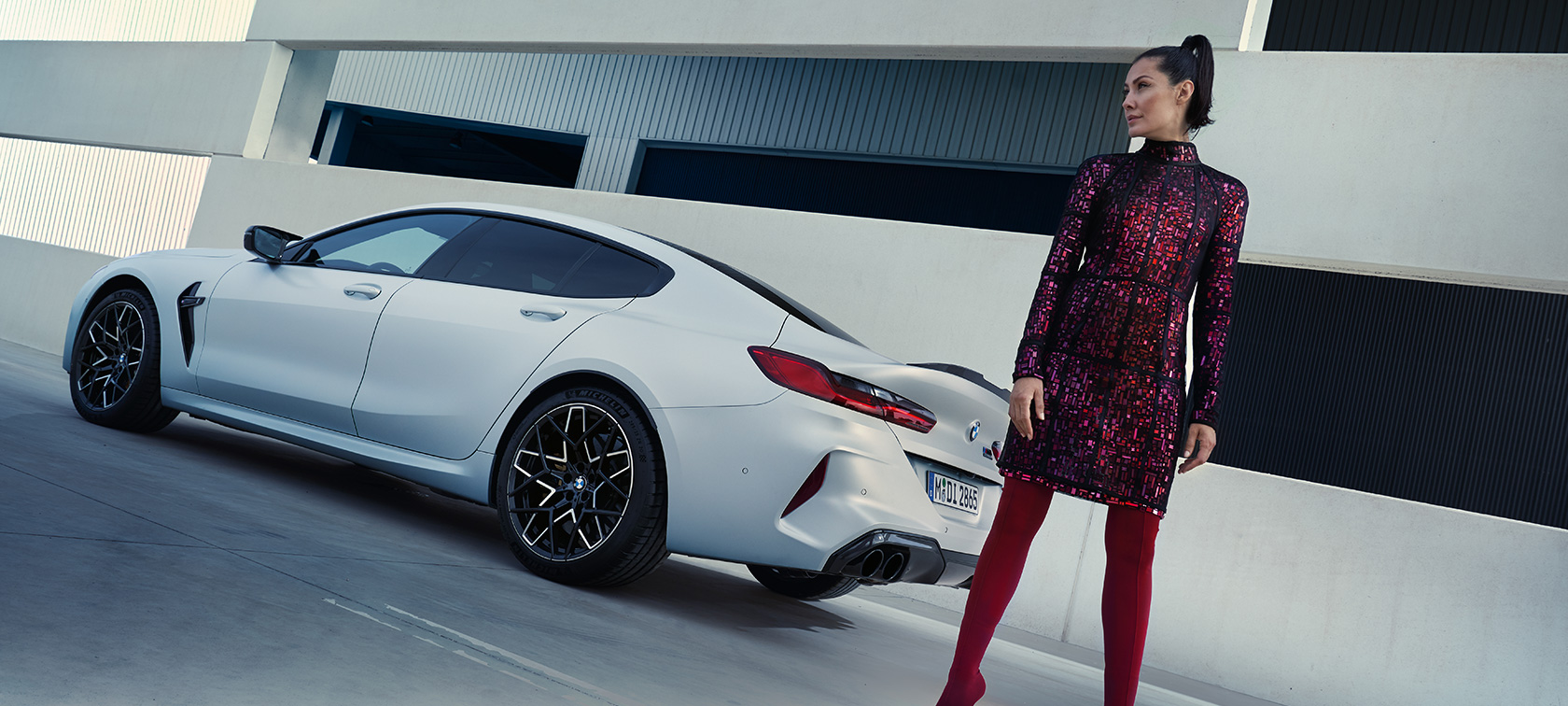 BMW M8 Competition Gran Coupé F93 arka çaprazdan görünüm BMW M8 Competition Gran Coupé F93 BMW Individual Metalik Beyaz/Frozen Brilliant otomobilin yanında duran kadın model ile arka çaprazdan görünüm
