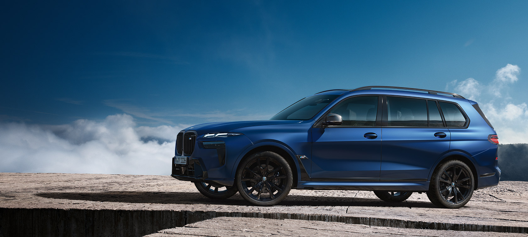 BMW X7 M60i G07 LCI, Metalik Mat Mavi/Marina Bay, dağ manzarası önünde hareketsiz halde yandan görünüm