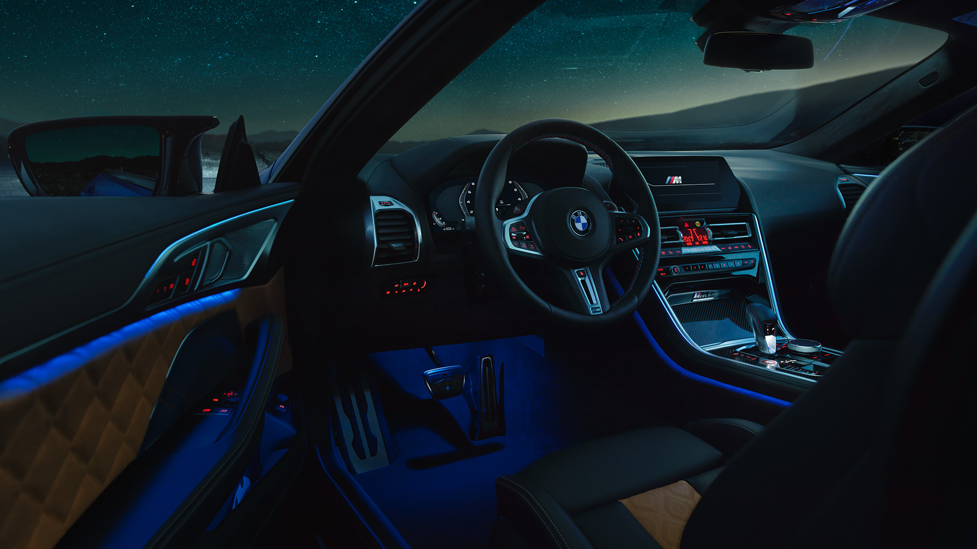 BMW M8 Competition Coupé’nin iç tasarımı. BMW M8 Competition Coupé, M deri direksiyon ve mavi ortam aydınlatmasıyla sürücü kapısı açık iç tasarımın gece görünümü.