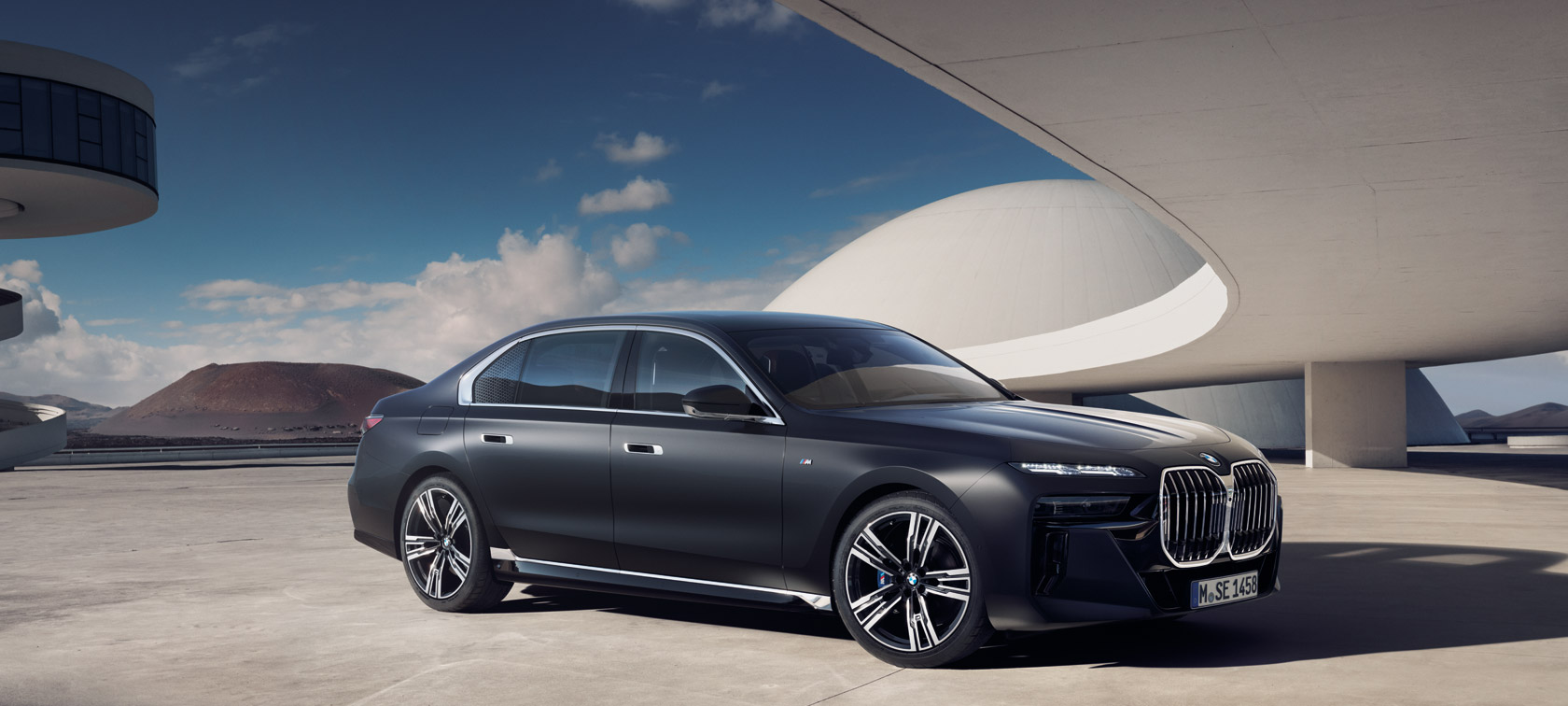 BMW 7 Serisi Sedan G70 ICE M Sport donanımı, Metalik Mat Koyu Gri, bina önünde hareketsiz halde ön çaprazdan görünüm