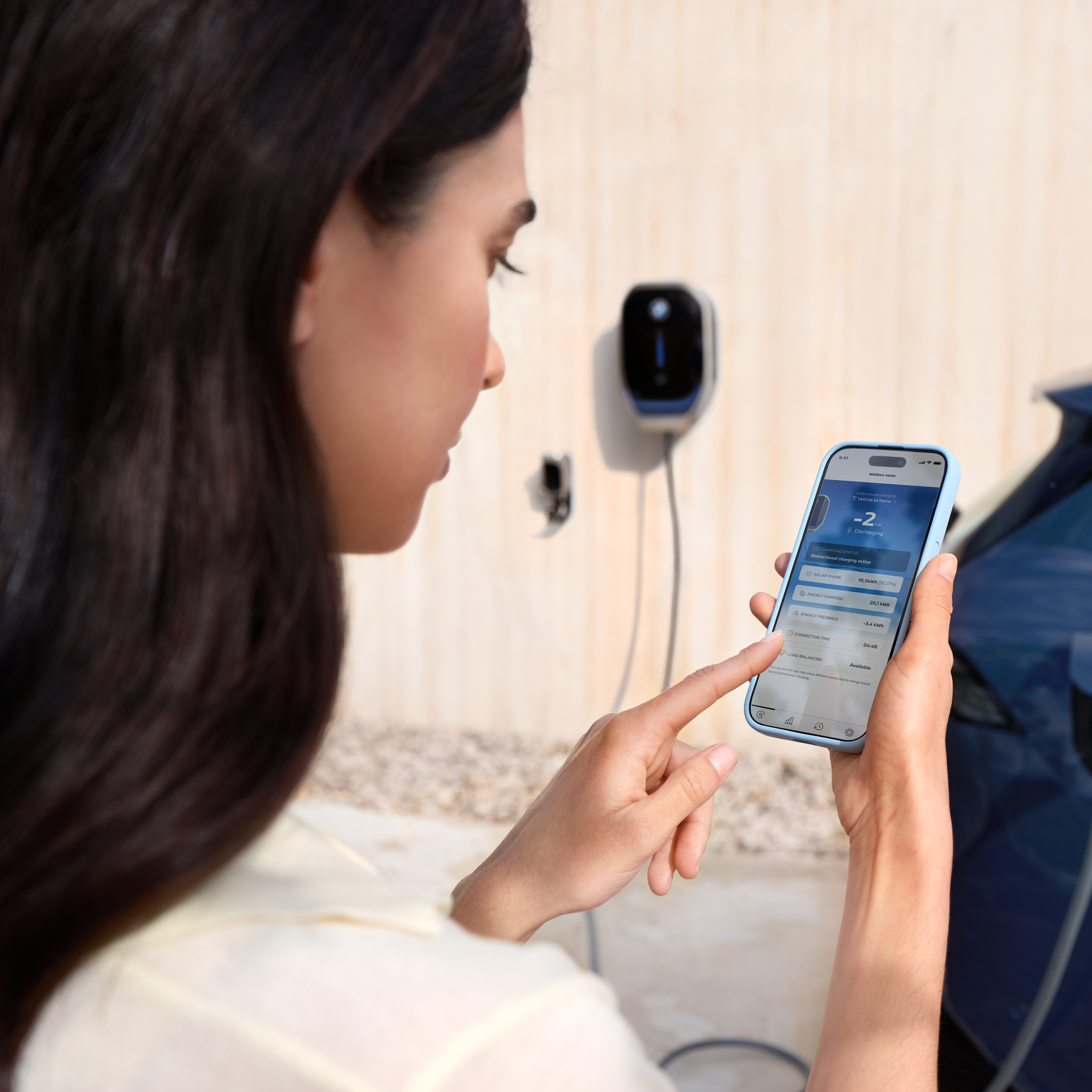 Bir kadın akıllı telefonuna bakıyor, arka planda wallbox görünüyor